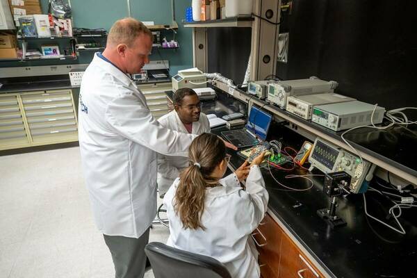 Three scientists in white labcoats examining a small device with colorful wires in a laboratory.